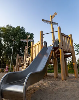 Ein hölzernes Spielplatzgebäude in Form eines Schiffes mit einem hohen Mast, Rutschen und Sand darunter. Im Hintergrund sind Bäume und ein klarer blauer Himmel zu sehen.