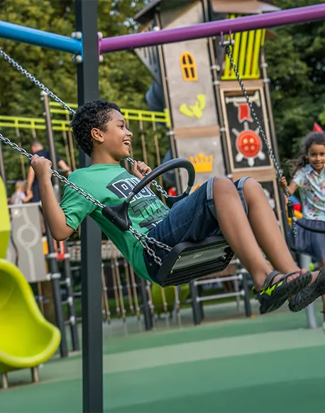 Ein lächelnder Junge in einem grünen Hemd und Shorts schwingt auf einer Spielplatzschaukel, mit bunten Spielgeräten und einem anderen Kind im Hintergrund.