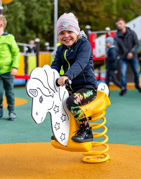 Ein lächelndes Kind, das eine gestreifte Mütze und bunte Leggings trägt, reitet auf einem weißen, gefederten Spielgerät in Form eines Pferdes. Andere Kinder und Erwachsene sind im Hintergrund zu sehen.