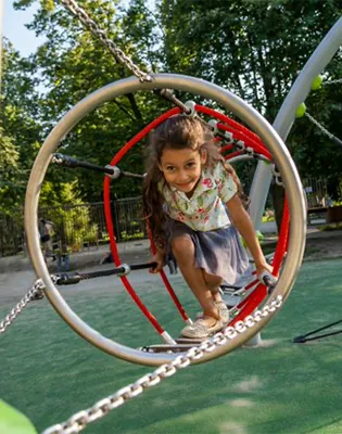 Ein junges Mädchen klettert lächelnd auf einen runden Spielplatz aus Metall und Seilen, während im Hintergrund Bäume und Grünflächen zu sehen sind.