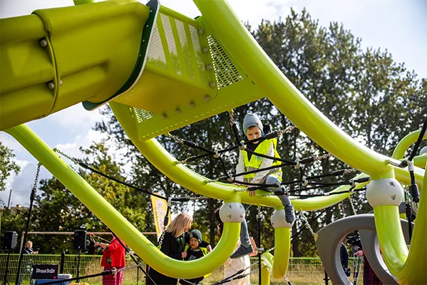 Ein Kind mit Hut und gelber Weste klettert auf ein großes, hellgrünes Klettergerüst auf einem Spielplatz im Freien, während Erwachsene und andere Kinder in der Nähe stehen. Im Hintergrund sind Bäume und ein bewölkter Himmel zu sehen.
