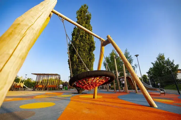 Ein moderner Spielplatz mit einer großen Korbschaukel auf einem Holzgestell, mit leuchtend orangefarbenem und blauem Gummibodenbelag, umgeben von Bäumen und Spielgeräten unter einem klaren blauen Himmel.