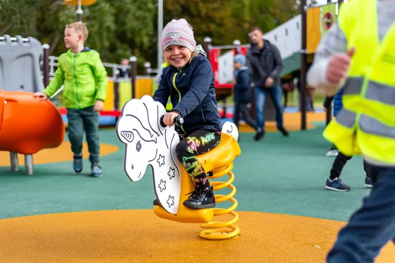 Ein lächelndes Kind mit einem gestreiften Hut reitet auf einer Federwippe in Form eines Pferdes auf einem Spielplatz, umgeben von anderen spielenden Kindern und bunten Spielgeräten.