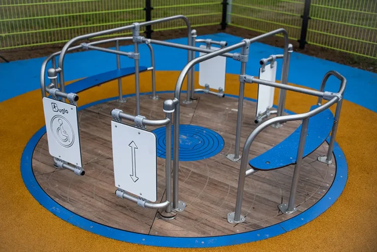 Ein barrierefreies Spielplatzkarussell mit Eingangstoren, Handläufen und Sitzgelegenheiten, das für Kinder mit Behinderungen konzipiert wurde. Die Oberfläche besteht aus Holz und Gummi und ist von einem Sicherheitszaun umgeben.